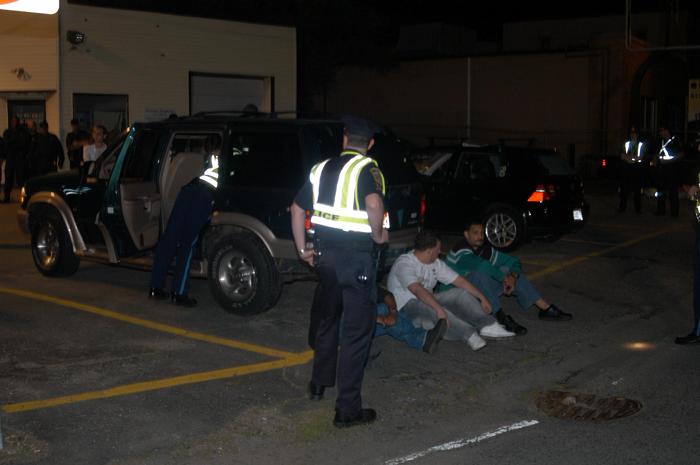 Police searching a car while people sit on the ground outside of it.